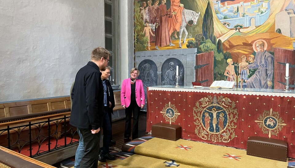Politikerne blir vist slitasjen på alterteppet, i bakgrunnen dem er alterveggen, malt av Hugo Lous Mohr i freskostilen, i 1932 , når dette maleriet var ferdig var det størst av sitt slag i Norge