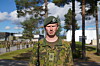 Soldat i kamuflasjeuniform og grønn beret står foran en militærleir, mens andre soldater står oppstilt i bakgrunnen.