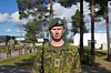 Soldat i kamuflasjeuniform og grønn beret står foran en militærleir, mens andre soldater står oppstilt i bakgrunnen.
